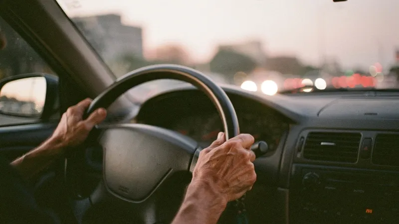 Las manos de un conductor adulto en el volante al atardecer, luces de la ciudad de fondo.